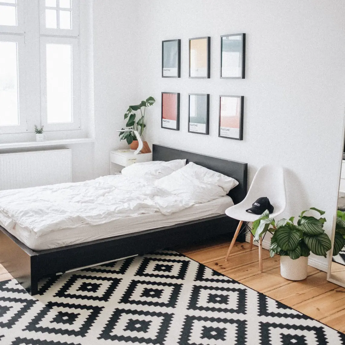 A bedroom features a black bed with white bedding and a geometric black and white rug. Above the bed, there are five framed pictures. A white chair holding a cap sits next to a potted plant. A window is on the left wall, while a mirror adorns the right wall. (This text has been generated by AI)