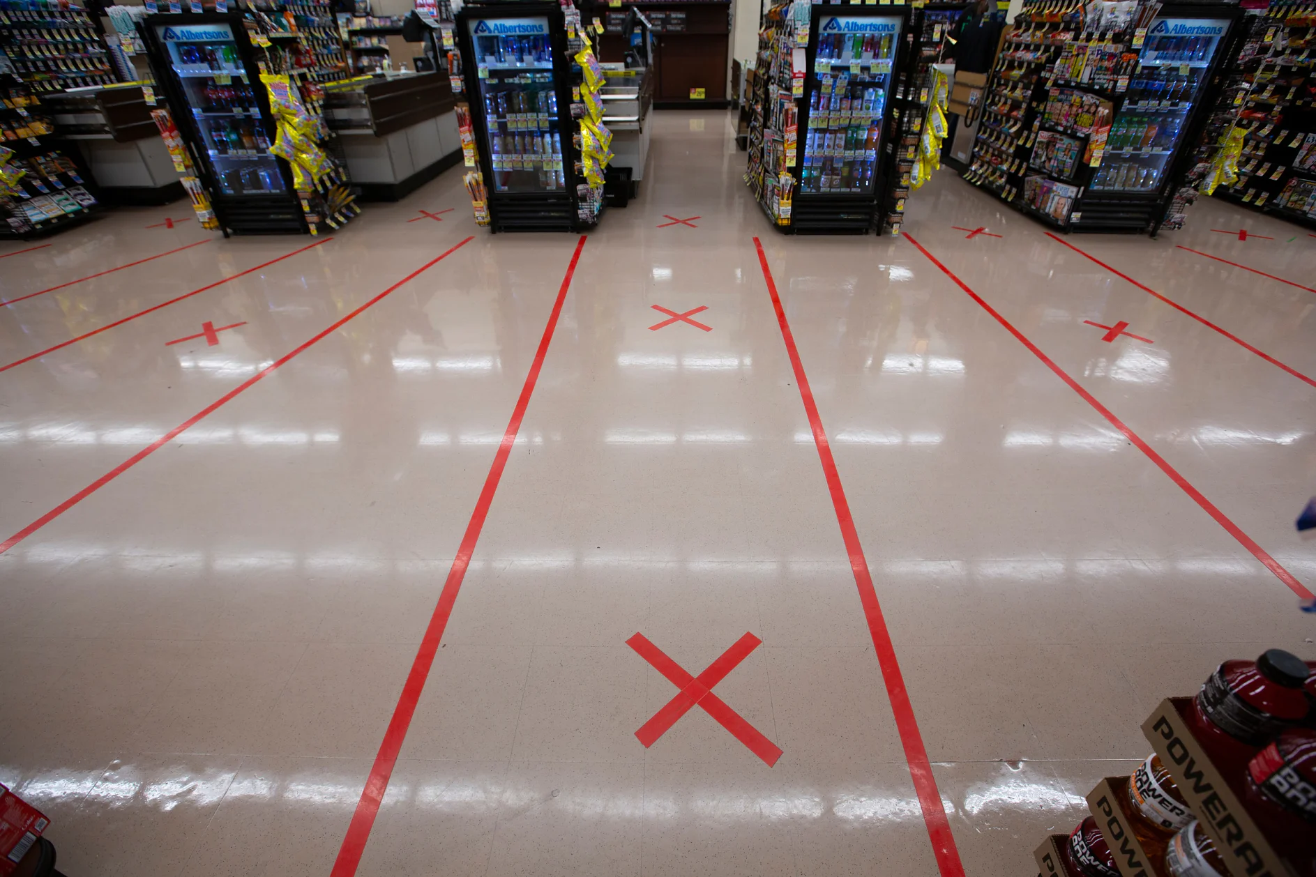 The image shows a grocery store aisle with red X marks and lines made of tesa tape on the floor, indicating social distancing measures. Shelves on both sides display various products, and coolers with drinks are visible ahead. (This text has been generated by AI)