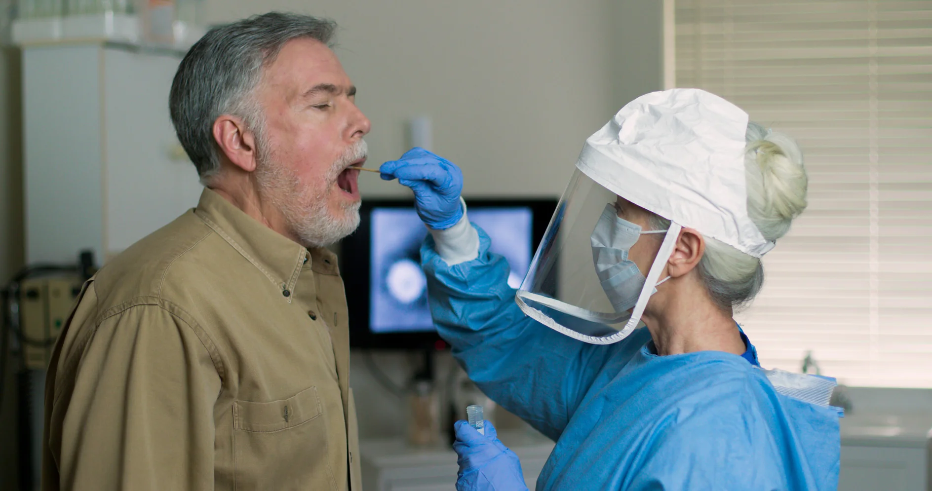 A medical professional in protective gear, including a face shield and mask, collects a nasal swab sample from an older man in a clinical setting. The man is standing with his mouth open, and a computer screen is visible in the background. (This text has been generated by AI)