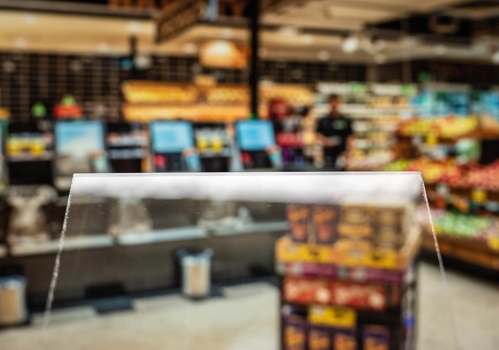 Blurred image of a supermarket interior showing various colorful products on shelves. In the foreground, a section of transparent protective glass is visible, while the background includes produce, cartons, and electronic checkout displays. (This text has been generated by AI)