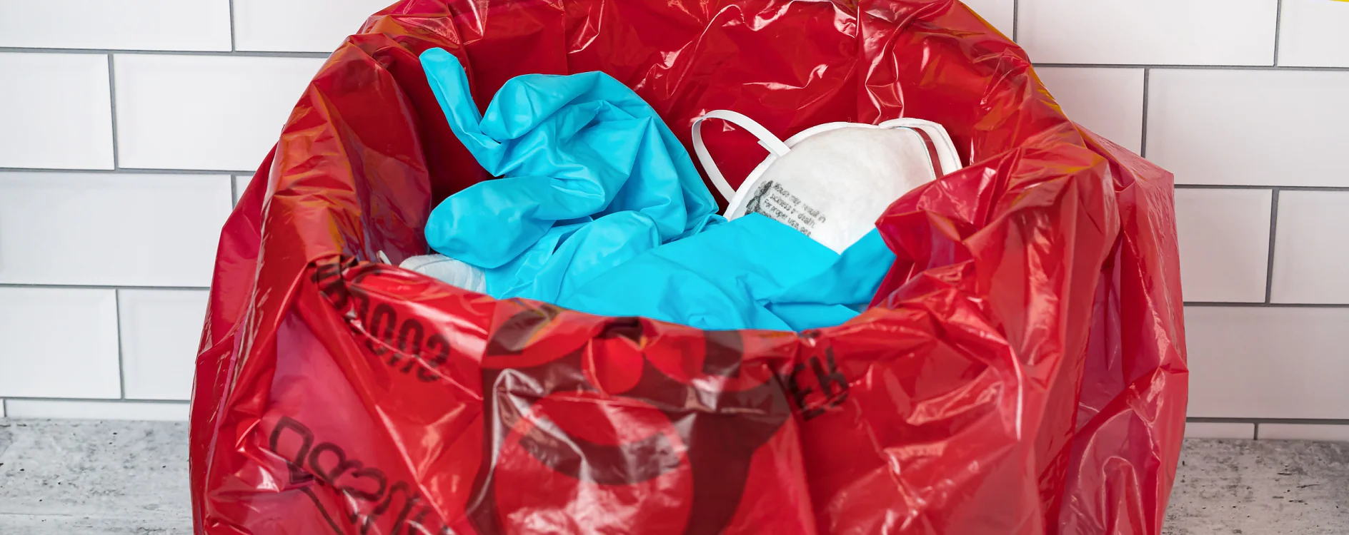 A red biohazard bag containing used protective equipment, including blue gloves and a white face mask, is placed on a gray surface in front of a tiled wall. All items are securely sealed with tesa tape. (This text has been generated by AI)