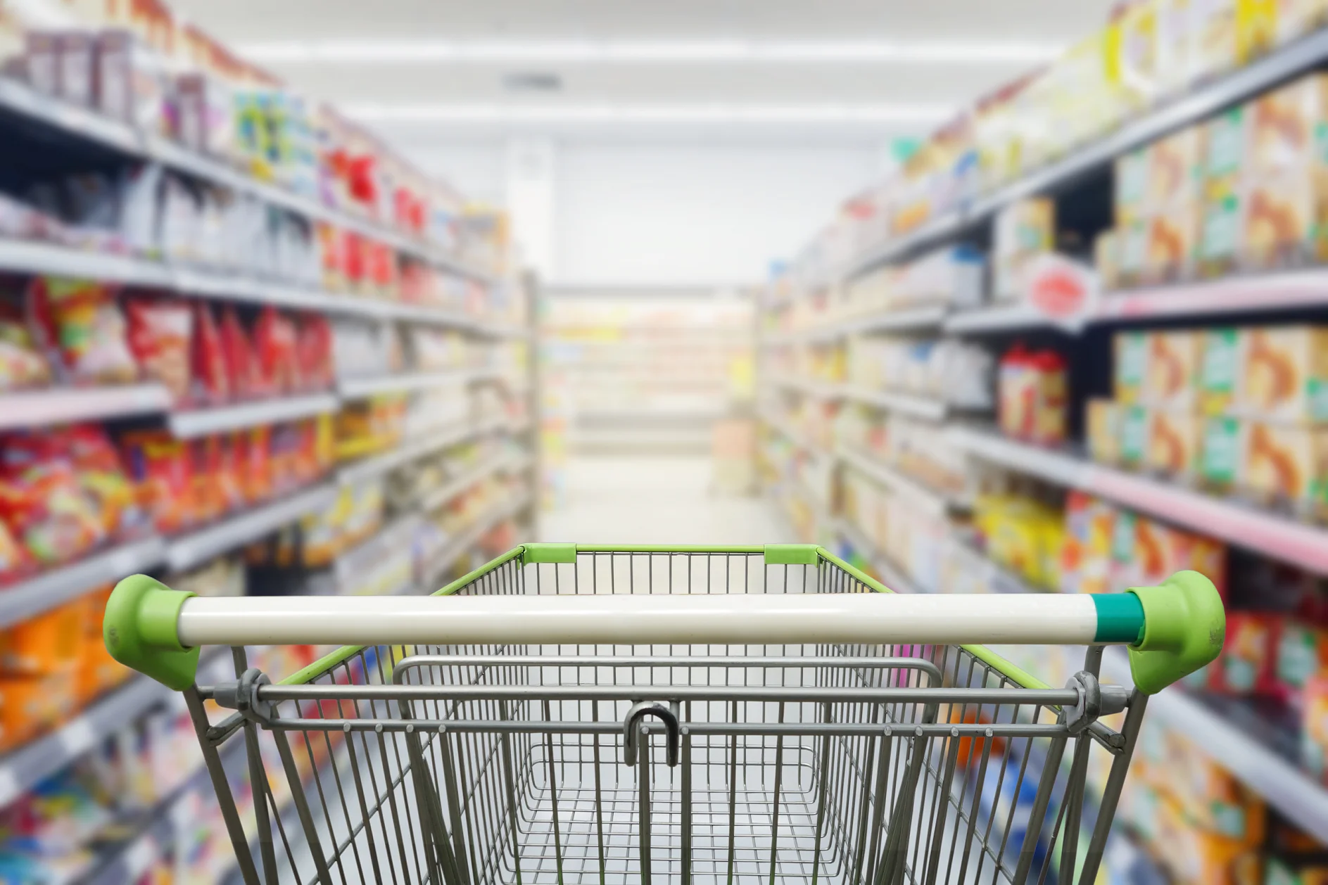 A shopping cart with green handles is positioned in an aisle of a grocery store. Shelves on both sides are stocked with various packaged goods, though the image is slightly blurred, emphasizing the tesa tape. (This text has been generated by AI)