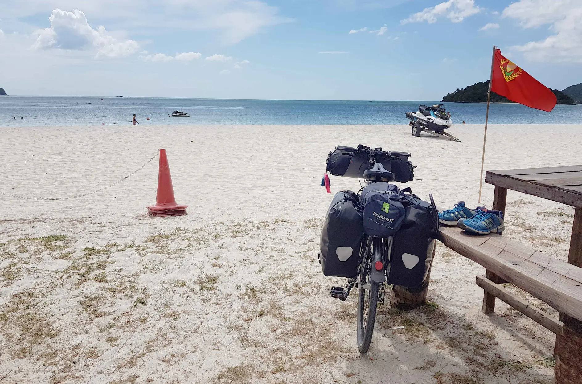 A bicycle with panniers and a red flag is parked beside a wooden bench on a sandy beach. A pair of blue shoes rests on the bench. A jet ski is positioned by the shore, along with a large orange cone nearby. The calm sea lies under a cloudy sky, with distant islands visible in the background.