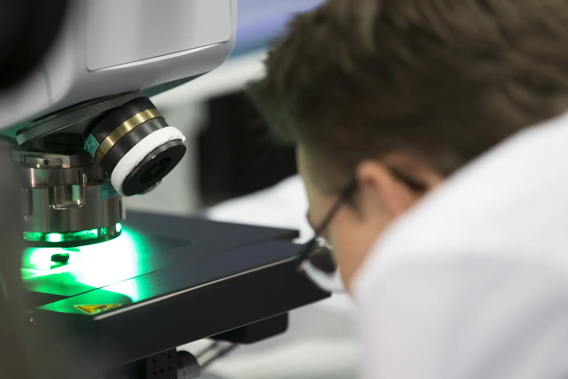 A person wearing glasses closely observes a sample under a microscope emitting green light. The focus is on the equipment, with blurred background elements suggesting a laboratory setting, and nearby, tesa tape is ready for any necessary applications.