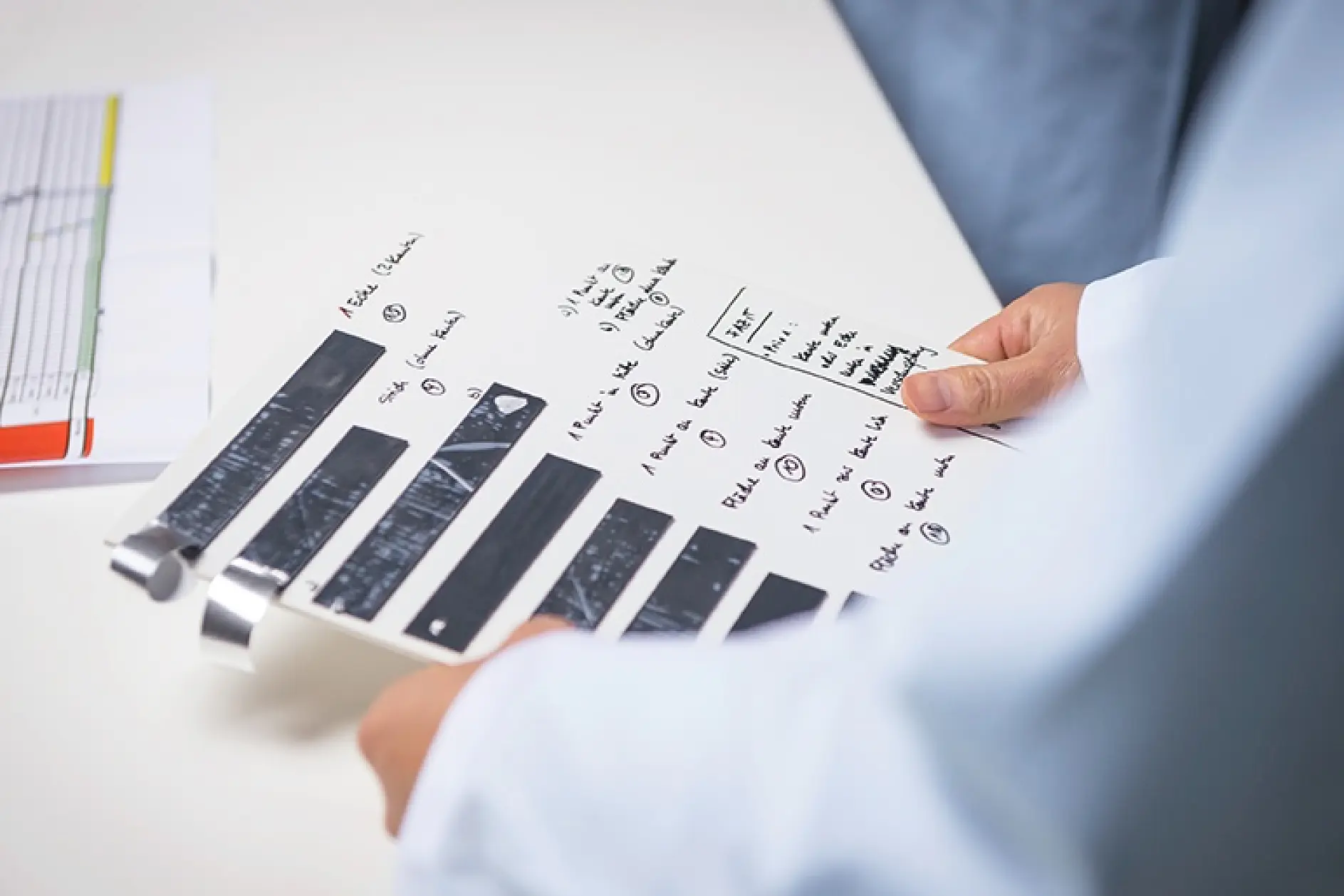 A person in a lab coat holds a sheet of paper with black strips of tesa tape and handwritten notes. The person is pictured from the shoulders down, with no visible face. A blurred chart is partially visible on the left side of the image.