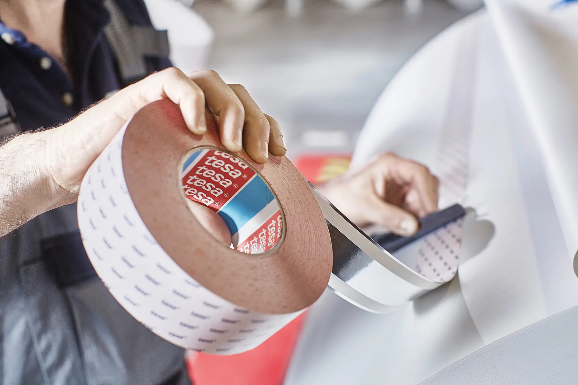 A person is holding a large roll of adhesive tesa tape with a red core labeled tesa. The tesa tape is being unwound, and another section of tesa tape is visible in the background. The person is wearing a dark shirt.