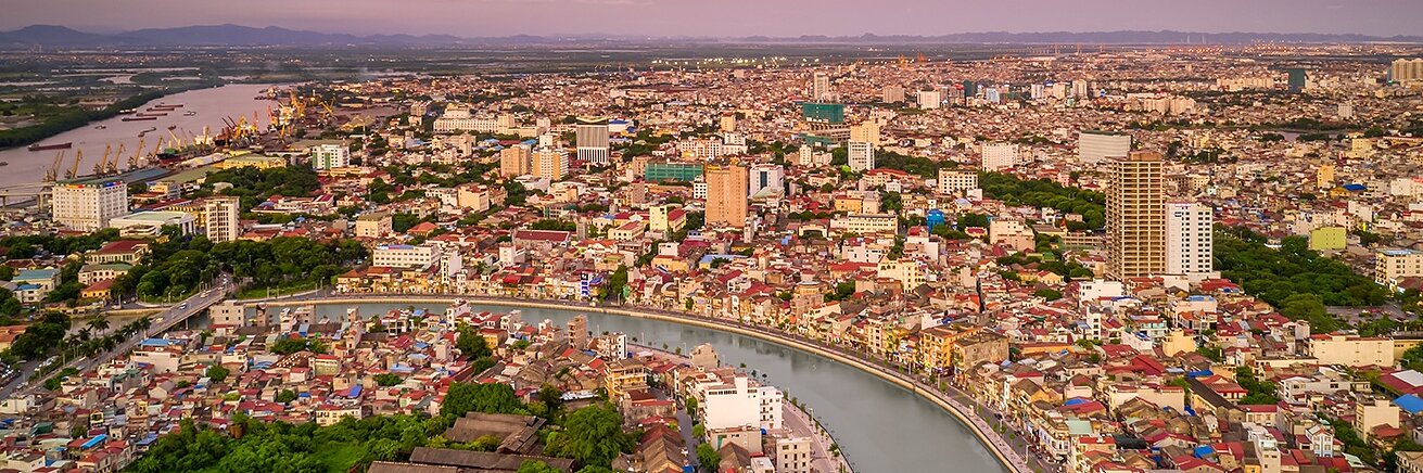 Aerial view of a cityscape during sunset with a river curving through the center. The skyline is dotted with buildings of varying heights. The sky displays hues of orange and pink, creating a warm ambiance.
