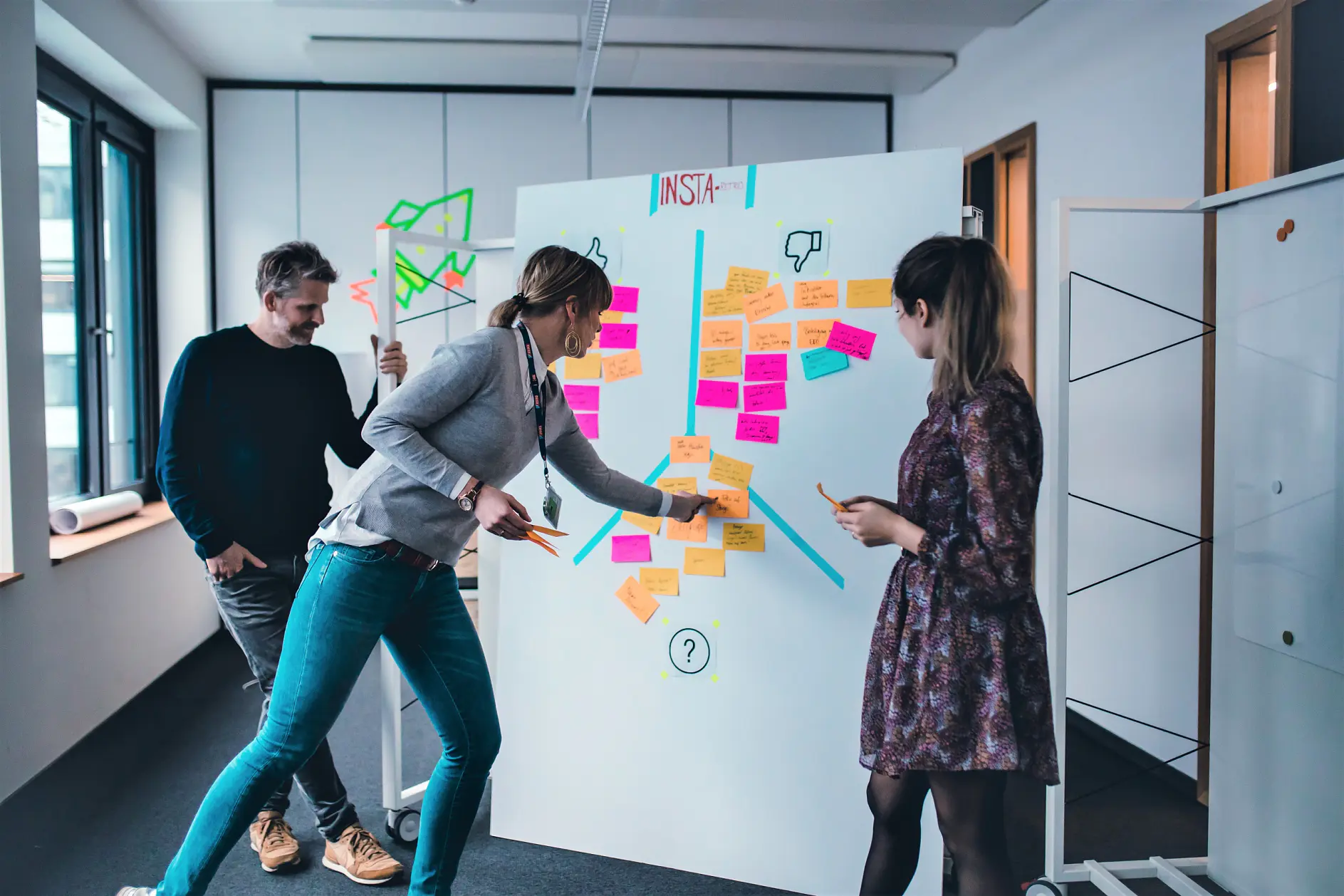 Three people in an office engage in a brainstorming session. They are placing and discussing colorful sticky notes on a whiteboard filled with various notes and symbols, using tesa tape to secure everything in place. The room is well-lit with windows on one side.