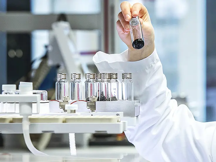 A scientist in a lab coat holds up a test tube containing a dark liquid. The background shows laboratory equipment and several other test tubes placed in a holder secured with tesa tape. The setting appears to be a laboratory environment.