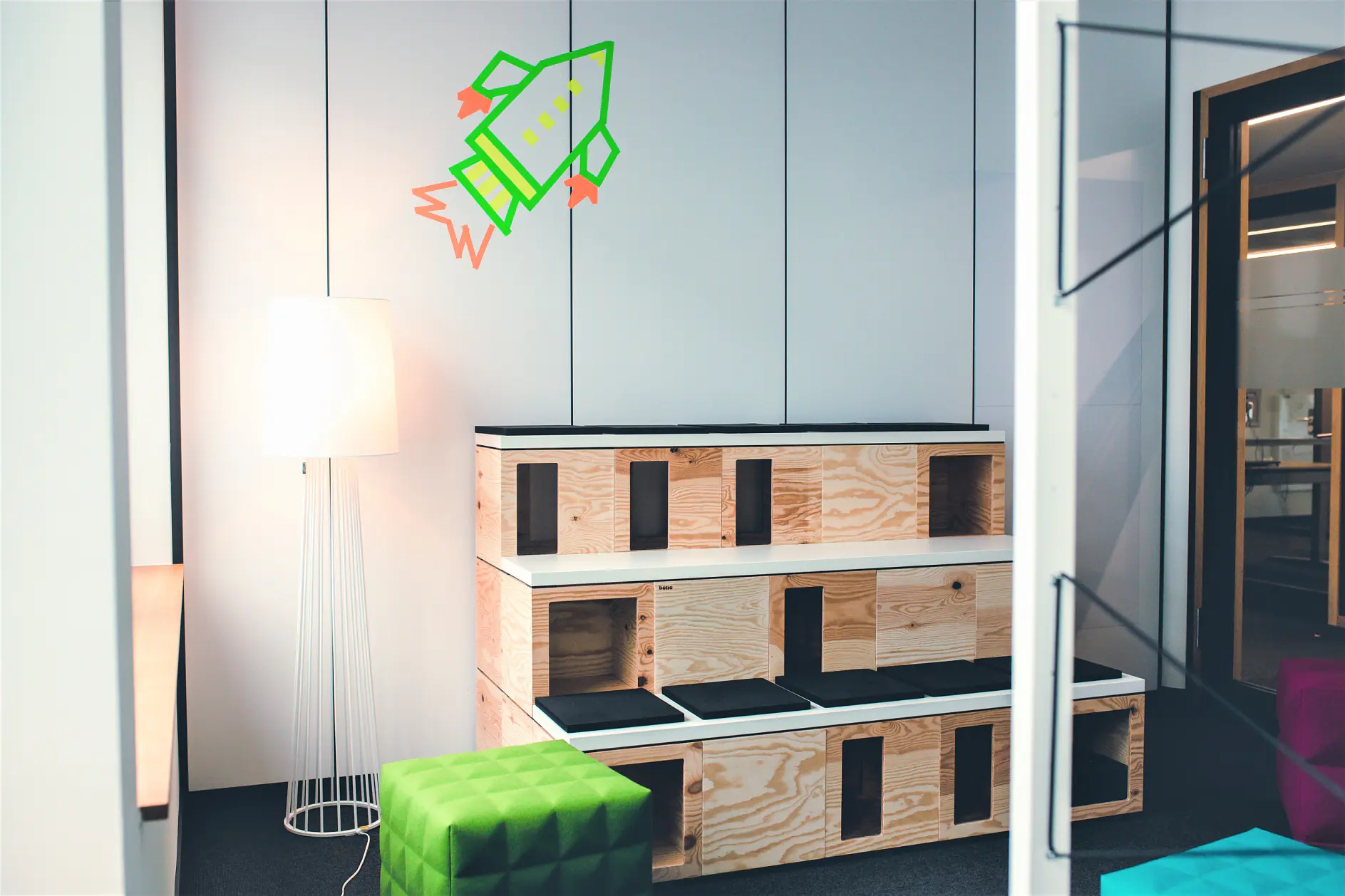 A modern interior features wooden modular seating against a white wall. A tall floor lamp is on the left, and a green rocket decal is secured with tesa tape on the wall above the seating. In front, theres a green quilted cube stool. The space is dominated by white and wood tones.