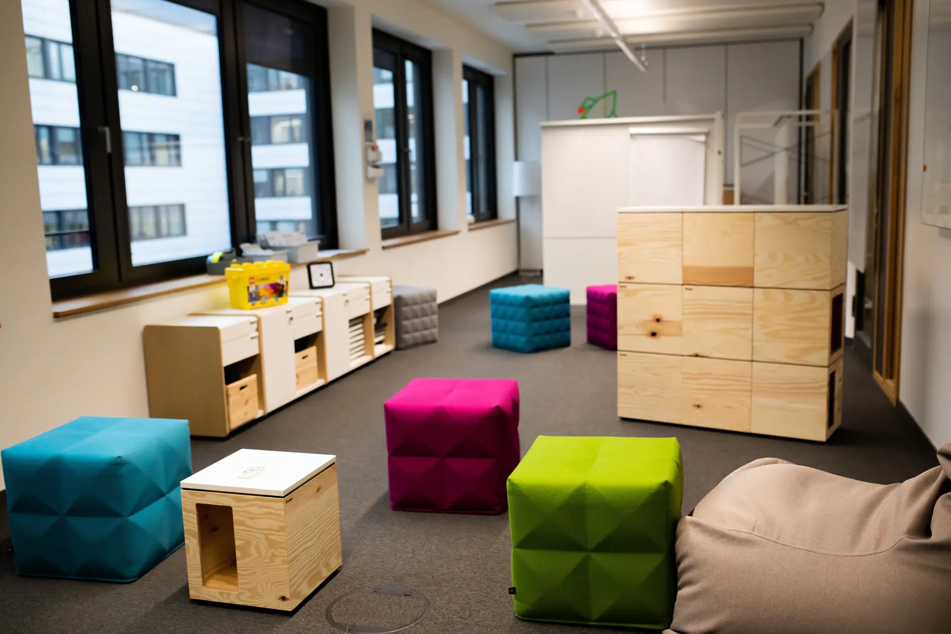 A modern childrens playroom with colorful cube seats in blue, pink, and green. Wooden storage units hold toys, and a whiteboard stands at the back. Large windows let in natural light. The room has a carpeted floor.