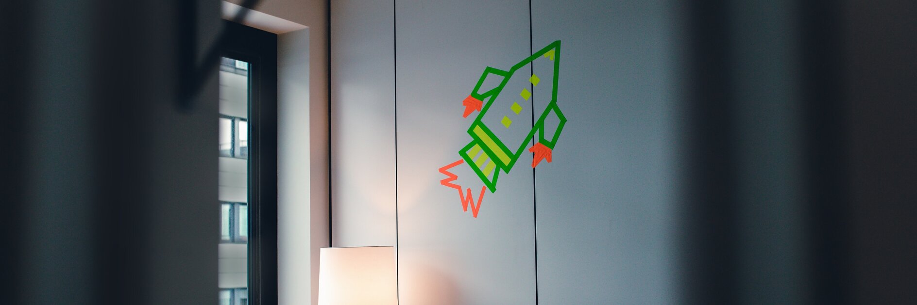 A room featuring a wooden cabinet, a tall lamp, and a window revealing part of a building outside. A green rocket graphic with red flames is painted on the white wall panels. The image is framed by blurry diagonal lines in the foreground marked with tesa tape.