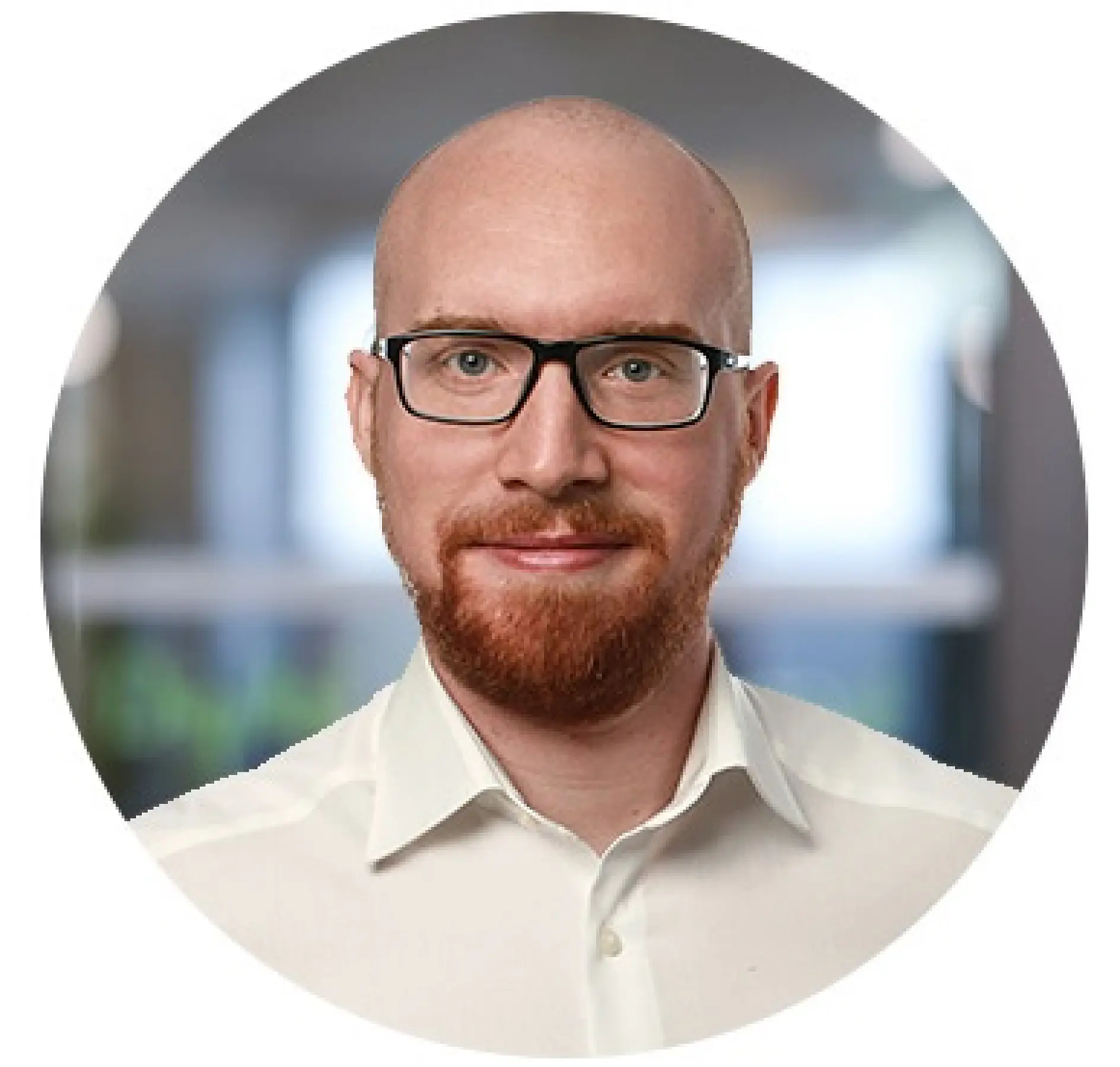 A bald man with a red beard and glasses is wearing a white collared shirt. The background is blurred, suggesting an indoor setting, possibly an office.
