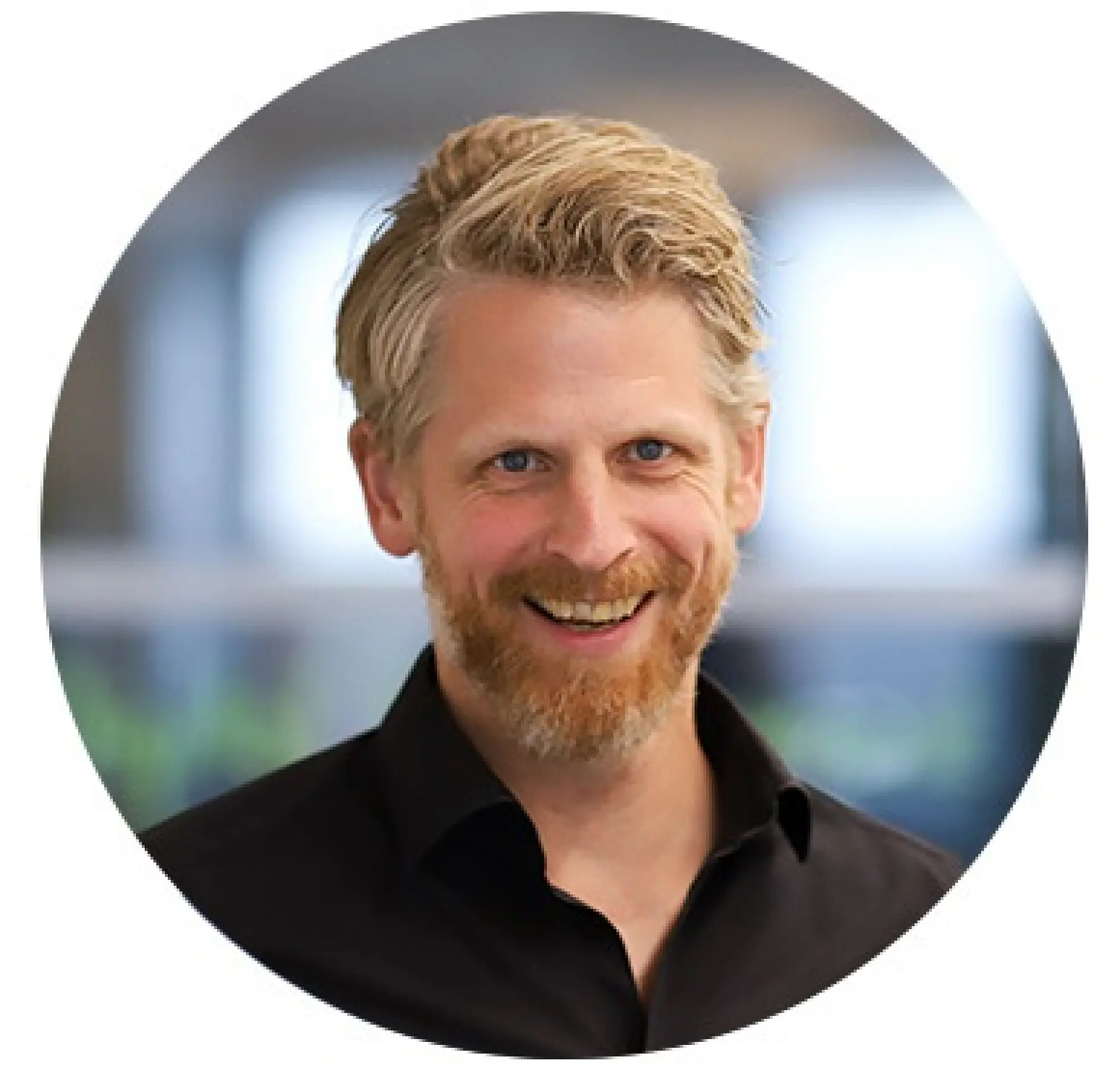 A man with short, wavy blond hair and a beard smiles at the camera. He is wearing a black shirt and is set against a blurred background of what appears to be an office environment. The image is circular, featuring various uses of tesa tape in the setting.