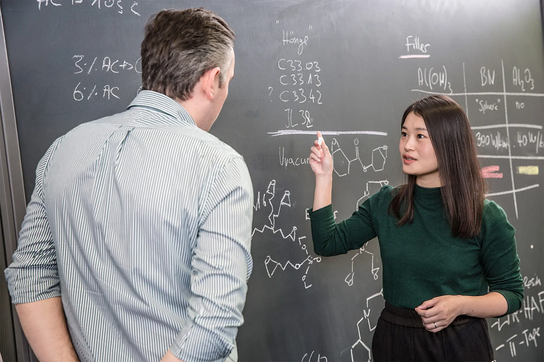 A woman in a green sweater explains scientific equations on a chalkboard to a man in a striped shirt. The chalkboard displays chemical structures and formulas. The setting appears to be an academic or research environment.