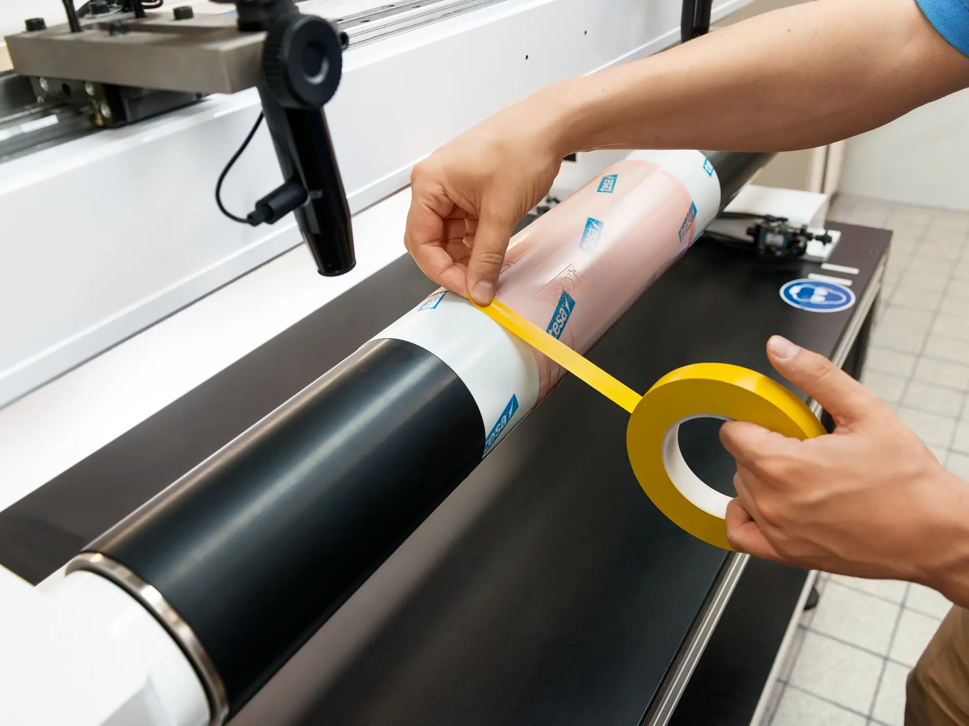 Person applying yellow tesa tape to a cylindrical object on a machine, surrounded by a workspace with a tiled floor. The object includes two sections, one black and one pink with blue logos.