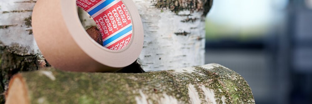 A roll of brown adhesive tesa tape is placed on a moss-covered log with other birch logs in the background. The scene is lit in natural light, emphasizing the texture of the tesa tape and the logs.