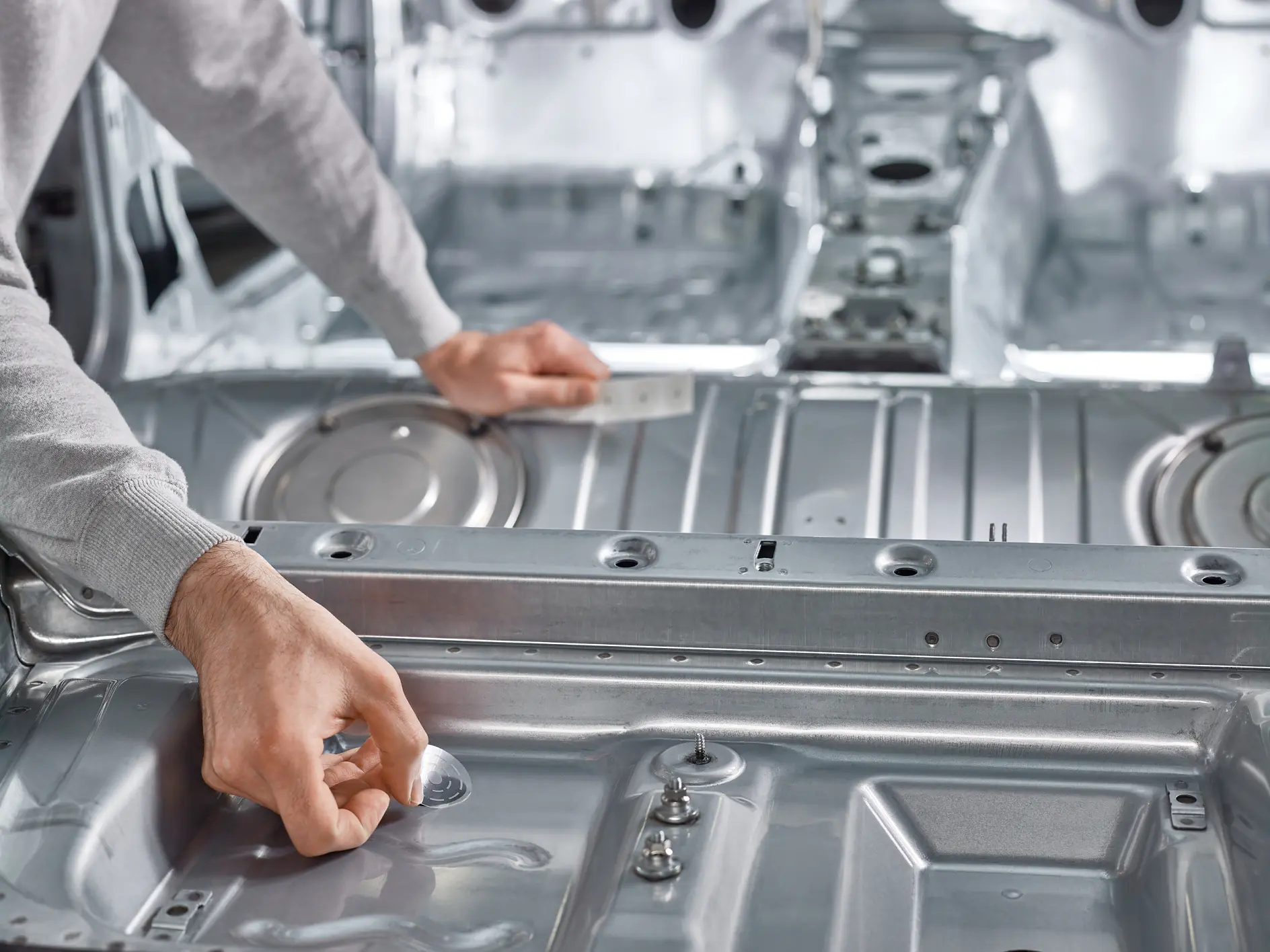 A person is assembling or inspecting a metallic automotive component. Their hands are adjusting or examining sections in the interior body frame of a car, using tesa tape. The background reveals a spacious, unpainted vehicle interior focused on the metal structure. (This text has been generated by AI)