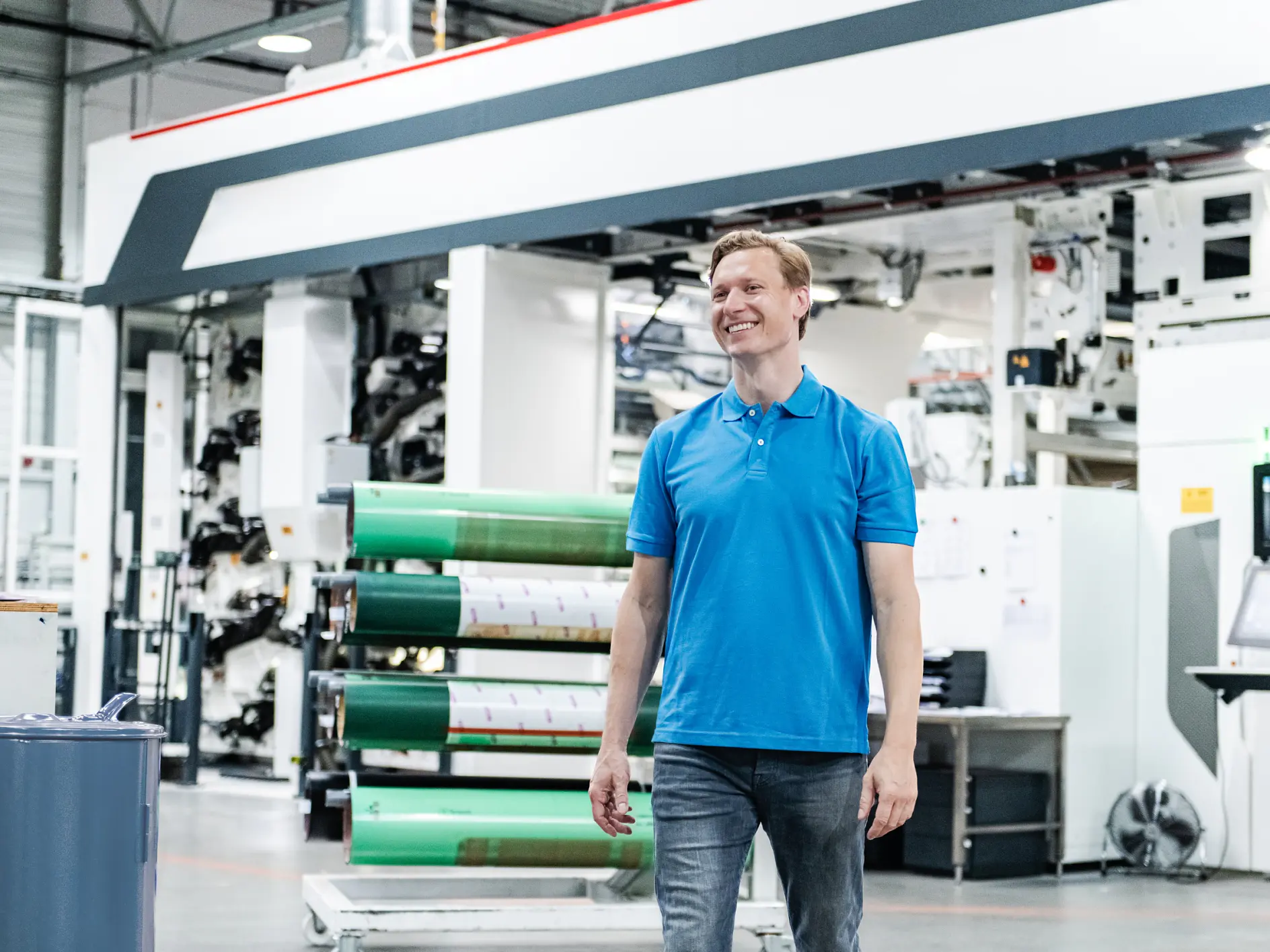 A man in a blue polo shirt and jeans is standing in a large industrial facility. Behind him, there is machinery and large rolls of tesa tape material. The setting appears to be a printing or manufacturing plant. (This text has been generated by AI)