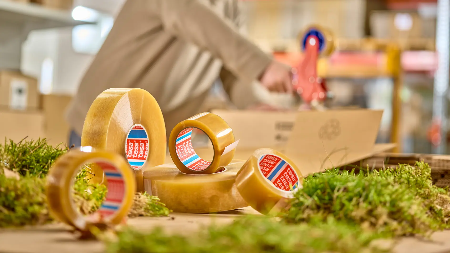 A person in a beige sweater is using a tesa tape dispenser in a warehouse setting. Several rolls of tesa tape are in the foreground, surrounded by patches of greenery. Shelves with boxes and pallets are visible in the background.
