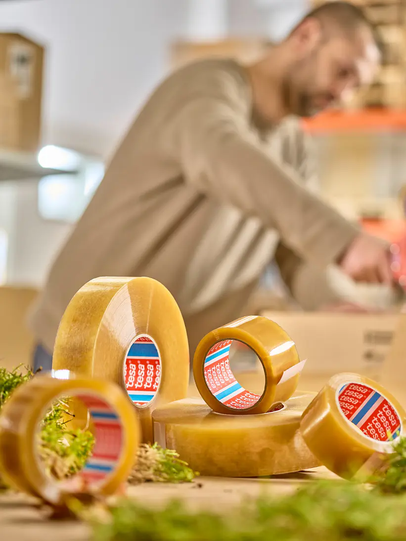 A person in a beige sweater is using a tesa tape dispenser in a warehouse setting. Several rolls of tesa tape are in the foreground, surrounded by patches of greenery. Shelves with boxes and pallets are visible in the background. (This text has been generated by AI)