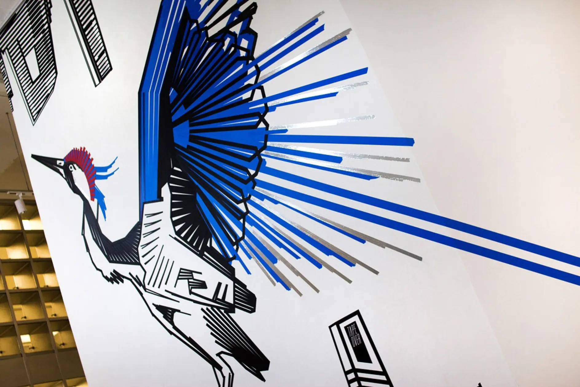 A mural of a stylized bird with exaggerated, geometric wings in black, blue, and gray lines adorns a white wall. The birds head features a red, rounded shape. The design incorporates angled details and abstract elements.