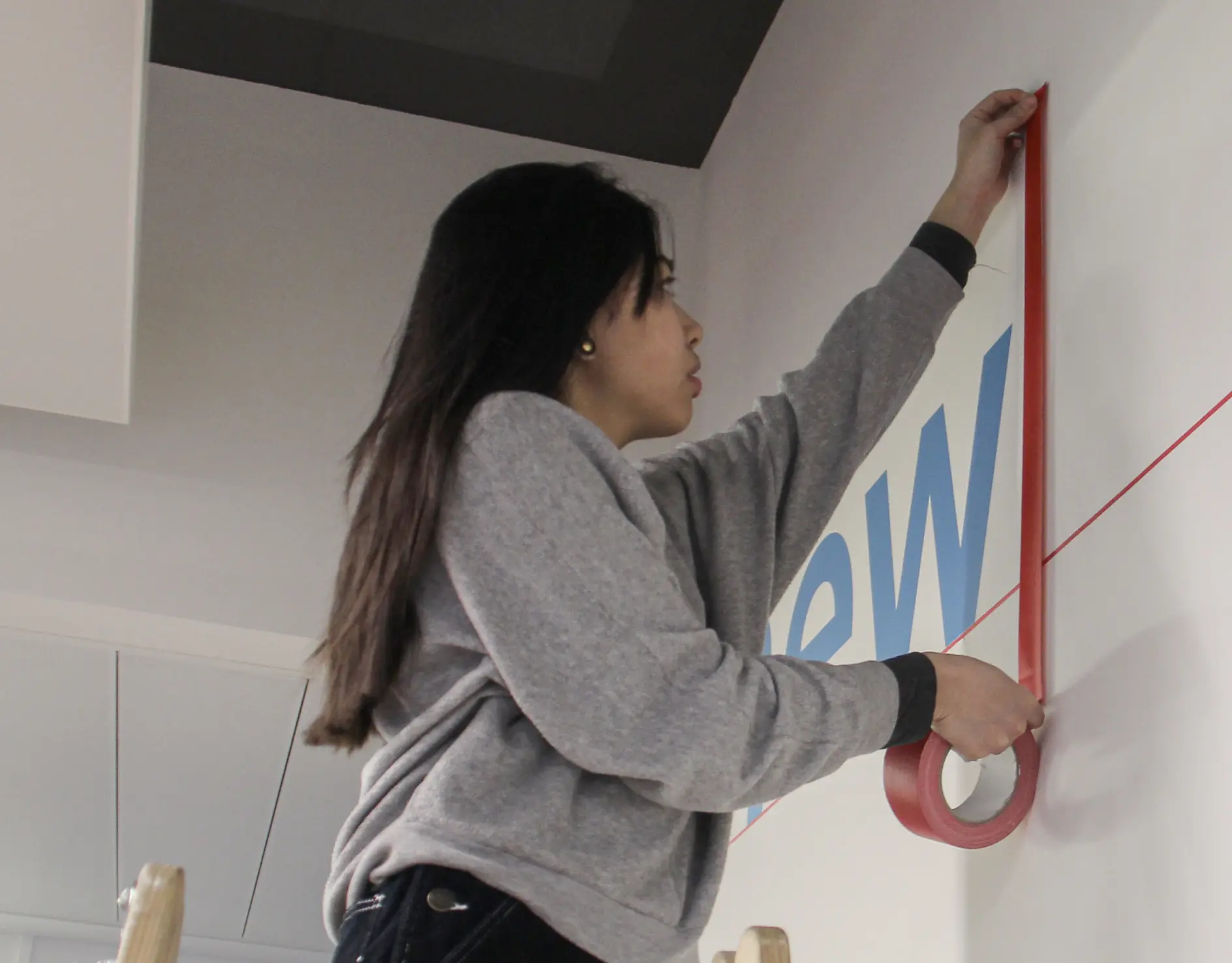 A person with long hair and a grey sweatshirt stands on a ladder, applying red tesa tape to a wall. The wall has partial blue lettering visible, with the letters ew showing. The setting appears to be indoors with a drop ceiling.