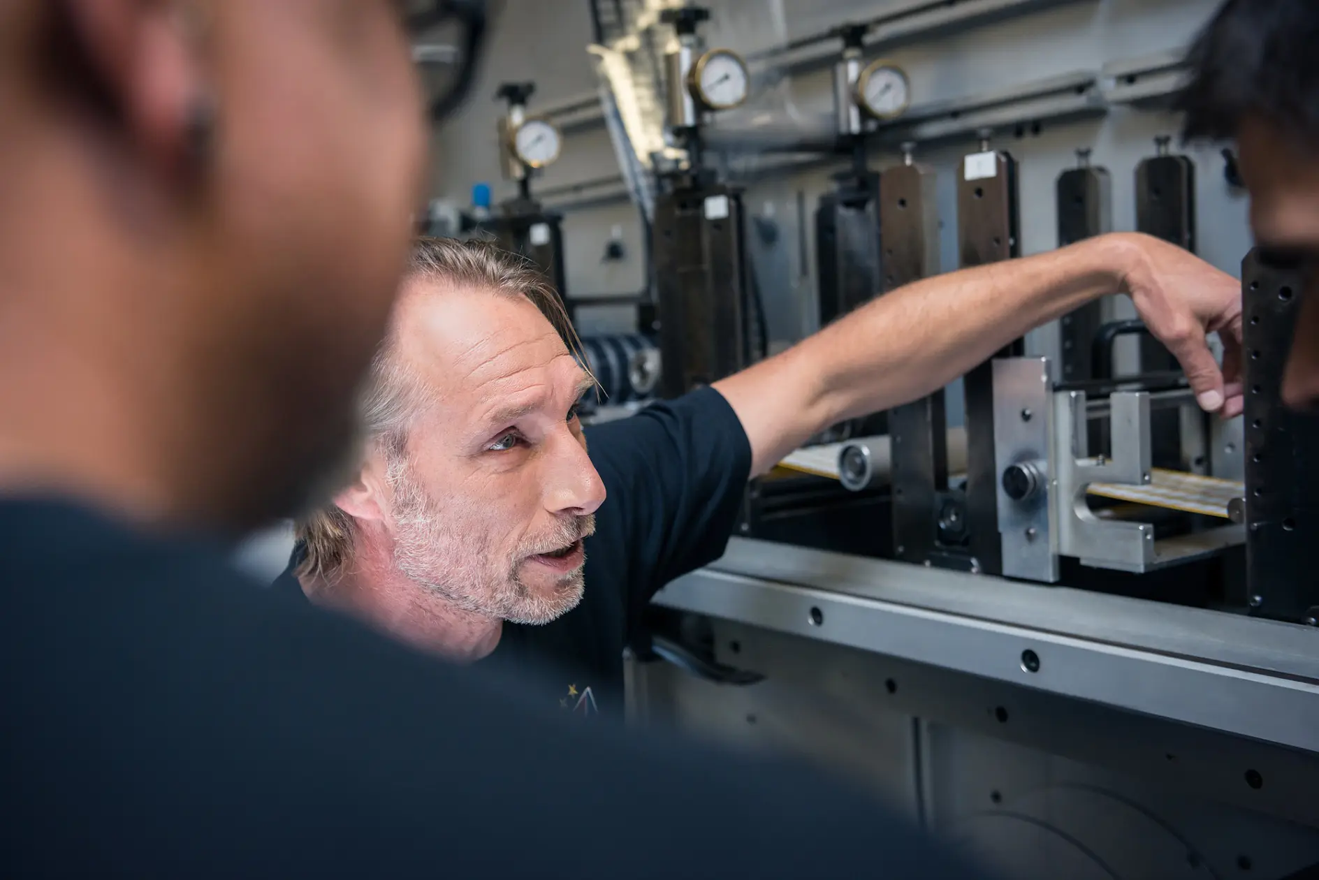 A person gestures towards machinery, explaining something to two others who are partially visible in the foreground. The setting appears to be an industrial or workshop environment with metal equipment and gauges, prominently featuring tesa tape.
