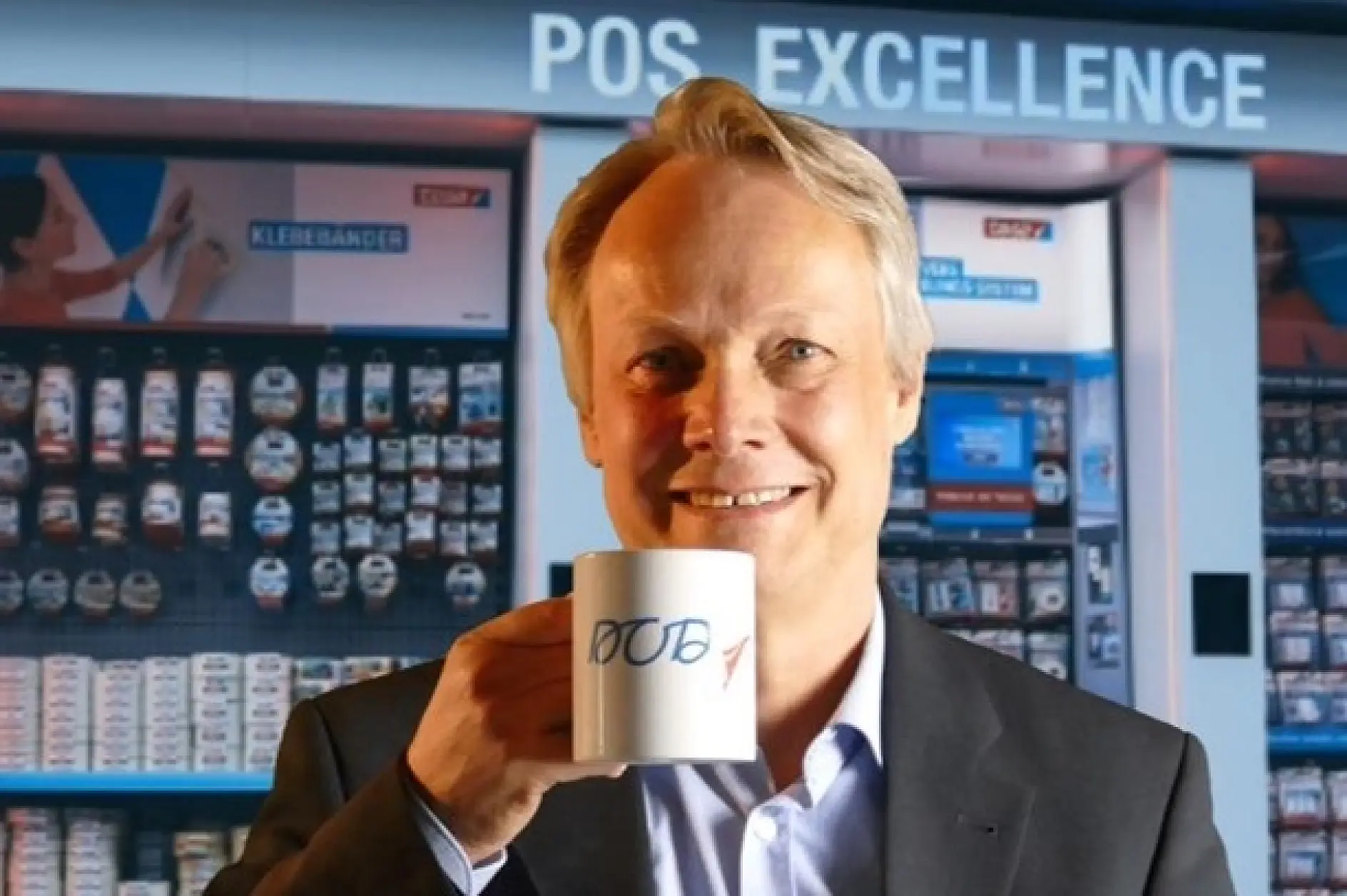 A person in a suit smiles while holding a white mug with JOB written on it. The background shows a retail display with the words POS EXCELLENCE above shelves stocked with various products, including tesa tape.