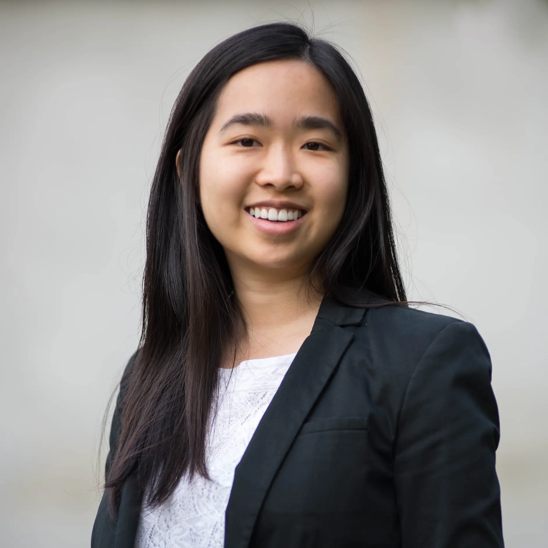 A person with long dark hair is smiling, wearing a black blazer over a white top. The background is neutral and blurred.