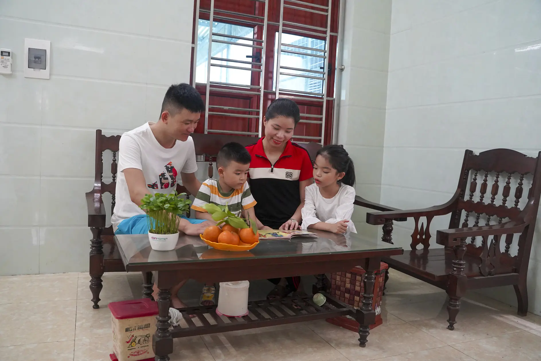 A family of four sits around a wooden table in a living room. The parents are smiling, while the young boy and girl focus on a book on the table. The room features light-colored walls and a window in the background.