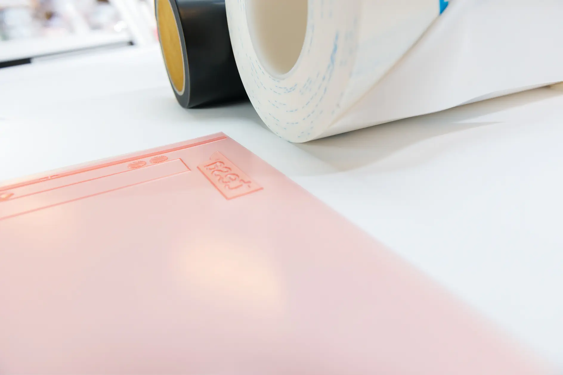 Close-up of a pink sheet with embossed text and symbols, placed on a white surface. Two rolls of tesa tape, one black and one white with blue patterns, are positioned in the background.