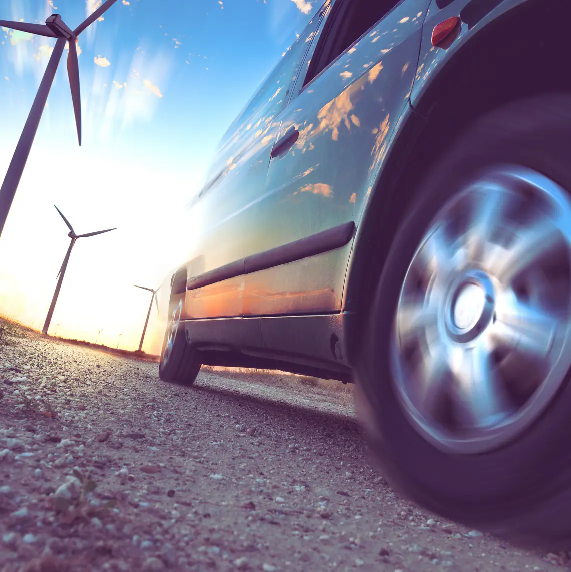 A vehicle is driving on a dirt road with a focus on its spinning wheel. Wind turbines are visible in the background against a sunset sky, casting reflections on the vehicles surface. The scene conveys a sense of motion and sustainable energy.