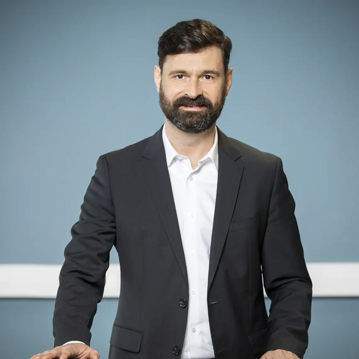 A man with dark hair and a beard is standing, wearing a black suit with a white shirt. He has both hands placed on a surface in front of him and is looking forward. The background is a muted blue with a white horizontal strip. On the table, you can see several rolls of tesa tape, neatly arranged next to some office supplies.