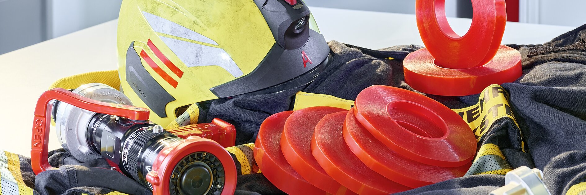 A firefighters helmet and hose are displayed with several red sealing rings on a yellow and black fabric. In the background, a fire extinguisher is visible through an open door.