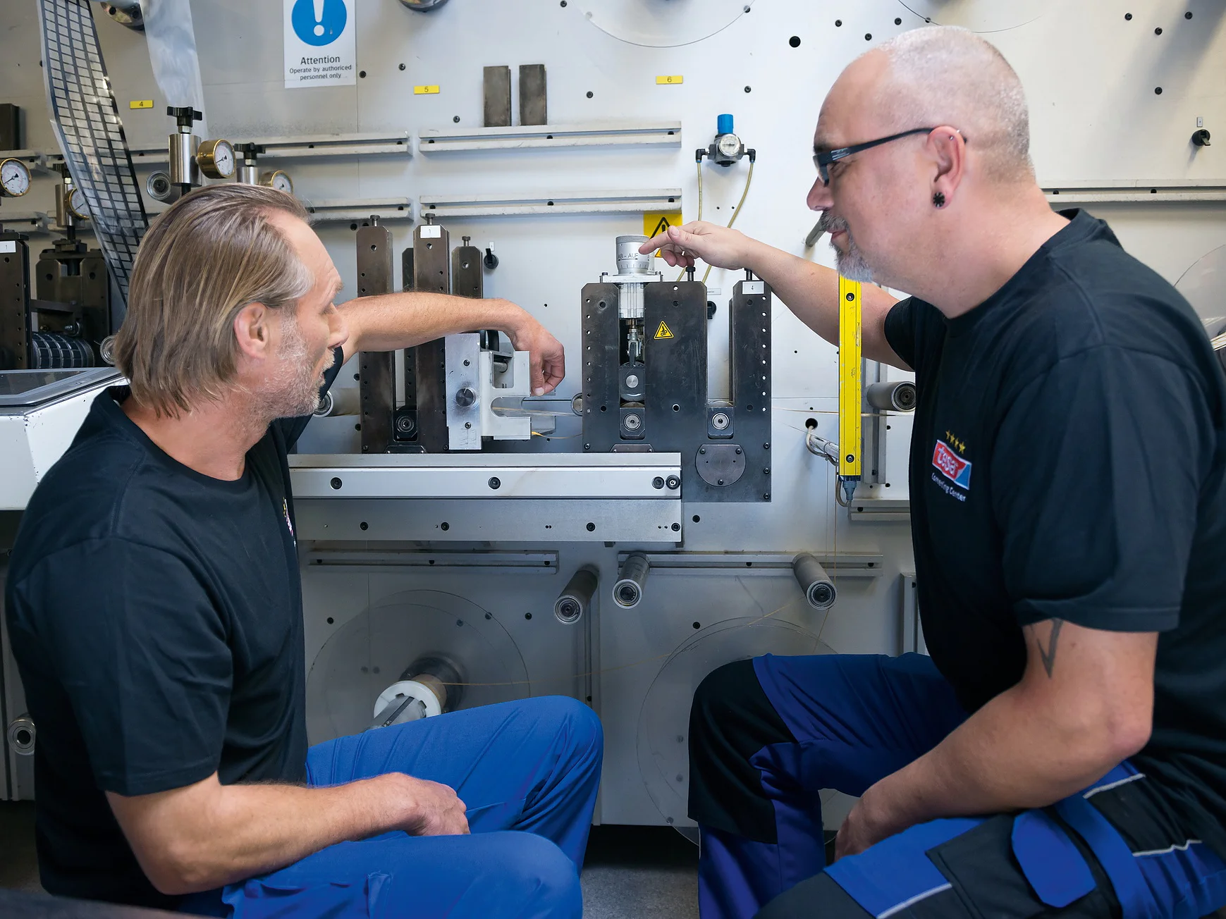 Two men are kneeling in front of industrial machinery, discussing its components. Both are wearing black shirts and blue pants. One man points at a part wrapped with tesa tape, while the other looks on attentively. The setting appears to be a workshop or factory. (This text has been generated by AI)