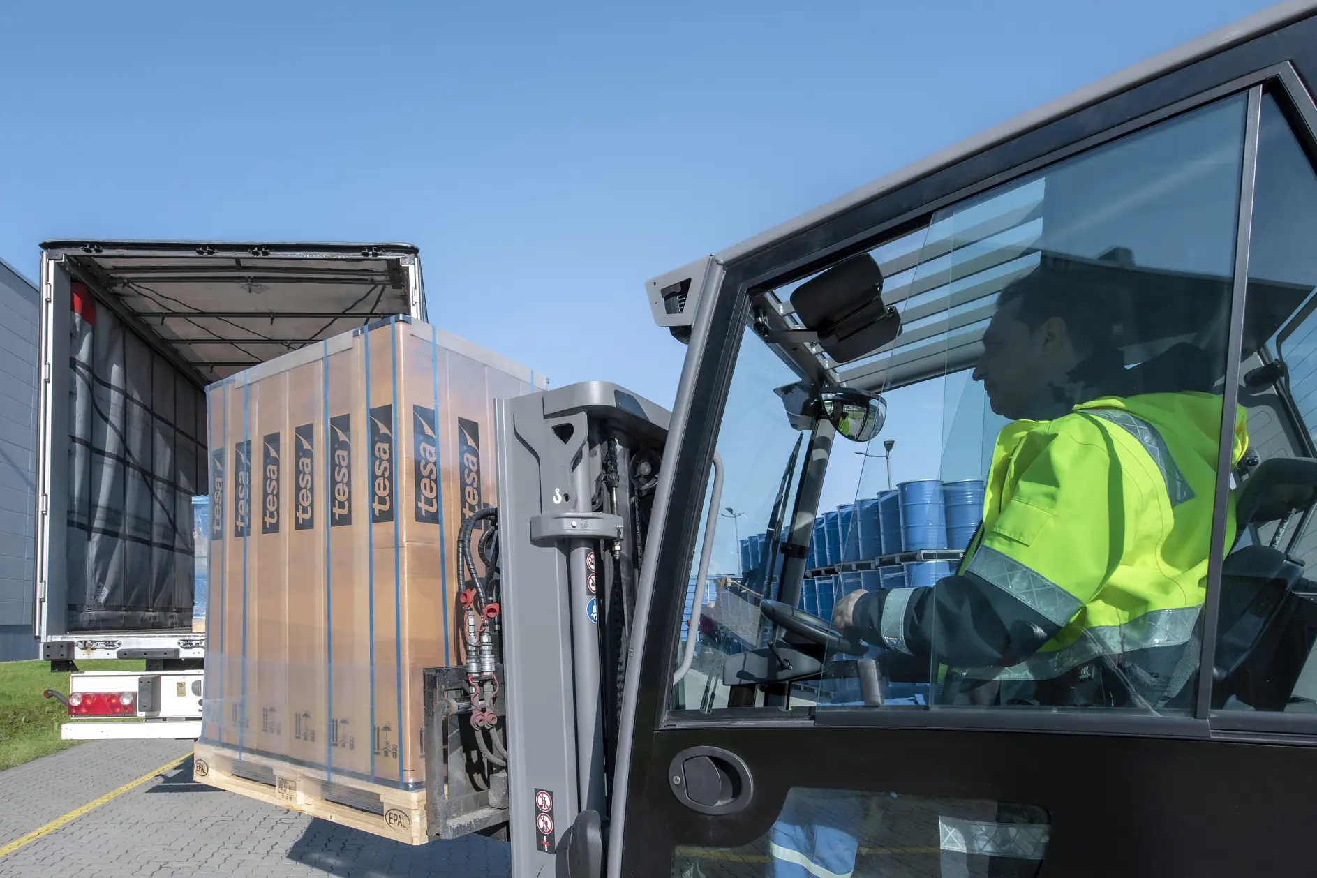 A forklift operator wearing a fluorescent yellow jacket loads a pallet of wrapped goods labeled Fessi into the back of a partially open truck. Blue barrels are visible in the truck. The scene takes place outdoors on a clear day, and everything is securely fastened with tesa tape.