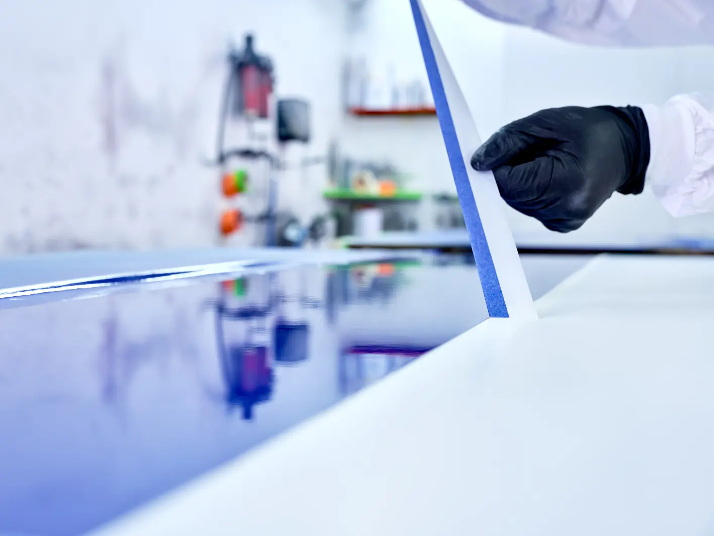 A person wearing black gloves applies blue tesa tape on a glossy blue surface. The background is an out-of-focus workshop with tools and equipment on the walls and shelves. The scene appears to be in a professional or industrial setting. (This text has been generated by AI)