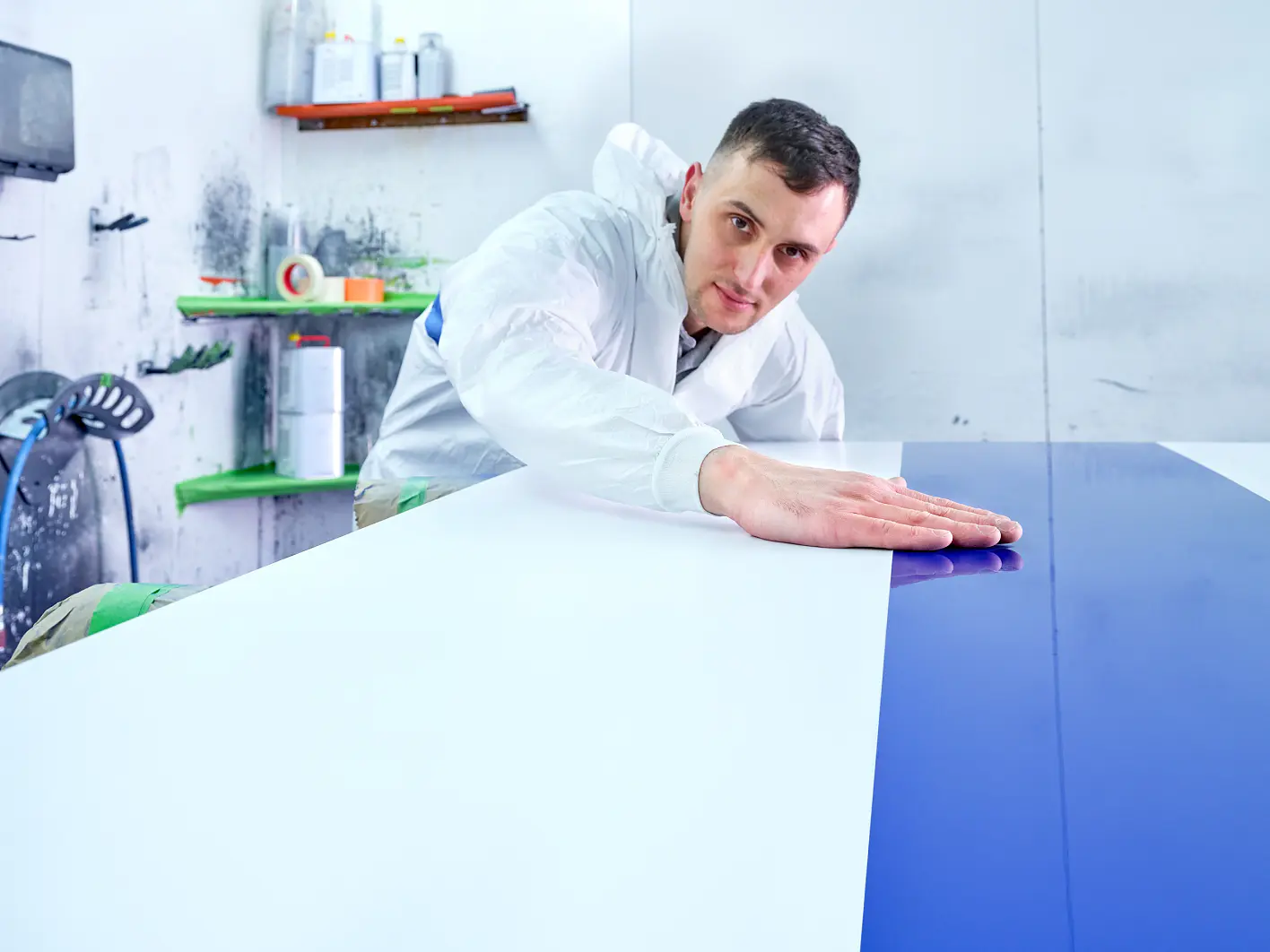 A person wearing a white protective suit is inspecting a large, smooth surface divided into white and blue sections. Their hand is placed on the surface. Shelves with various containers are visible in the background, marked by precise applications of tesa tape throughout. (This text has been generated by AI)