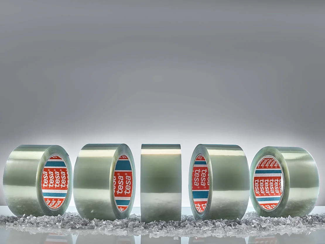 Five rolls of clear adhesive tesa tape are displayed vertically against a gray background. Small, scattered transparent granules are on the surface in front of the rolls. The tape cores are branded with red and blue labels. (This text has been generated by AI)