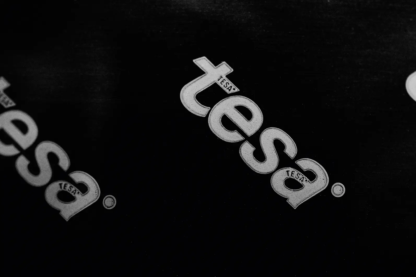 Black and white image showing multiple diagonal logos of the brand tesa tape. The logos are white with a small registered trademark symbol and letter T inside an oval next to each one, set against a dark background. (This text has been generated by AI)