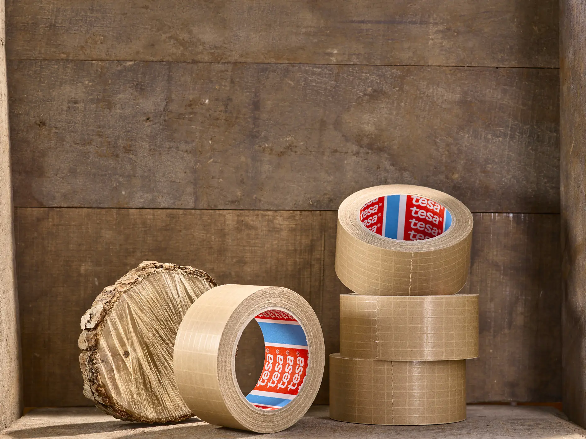 Several rolls of brown packing tesa tape are stacked and displayed on a wooden surface. A round piece of wood with a textured bark edge is placed beside the tesa tape rolls. (This text has been generated by AI)