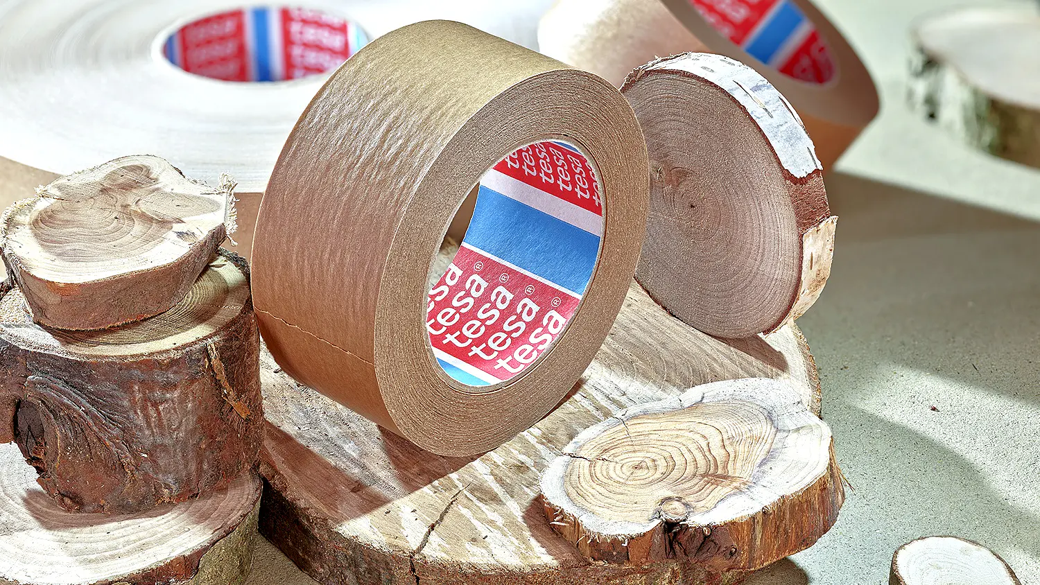A roll of brown tesa tape is placed on round wooden slices. The tesa tape features a red and blue label inside. The wooden slices vary in size and have visible tree rings, adding a natural element to the setup. (This text has been generated by AI)