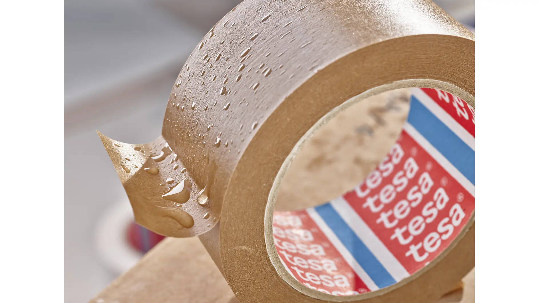 A close-up of a roll of brown adhesive tesa tape with water droplets on the surface. The inner side of the roll shows red, white, and blue branding with the word tesa. The tesa tape is partially unrolled against a blurred background. (This text has been generated by AI)
