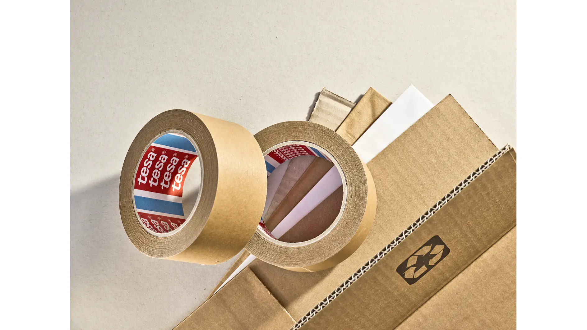Two rolls of brown packing tesa tape, with visible brand labels, rest on top of corrugated cardboard and white cardstock against a neutral background. A recycling symbol is printed on one of the cardboard pieces. (This text has been generated by AI)