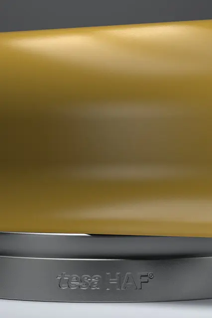 A large roll of yellow adhesive tesa tape is displayed on a gray base against a dark background. The inner side of the tesa tape roll has a red, blue, and white pattern. (This text has been generated by AI)