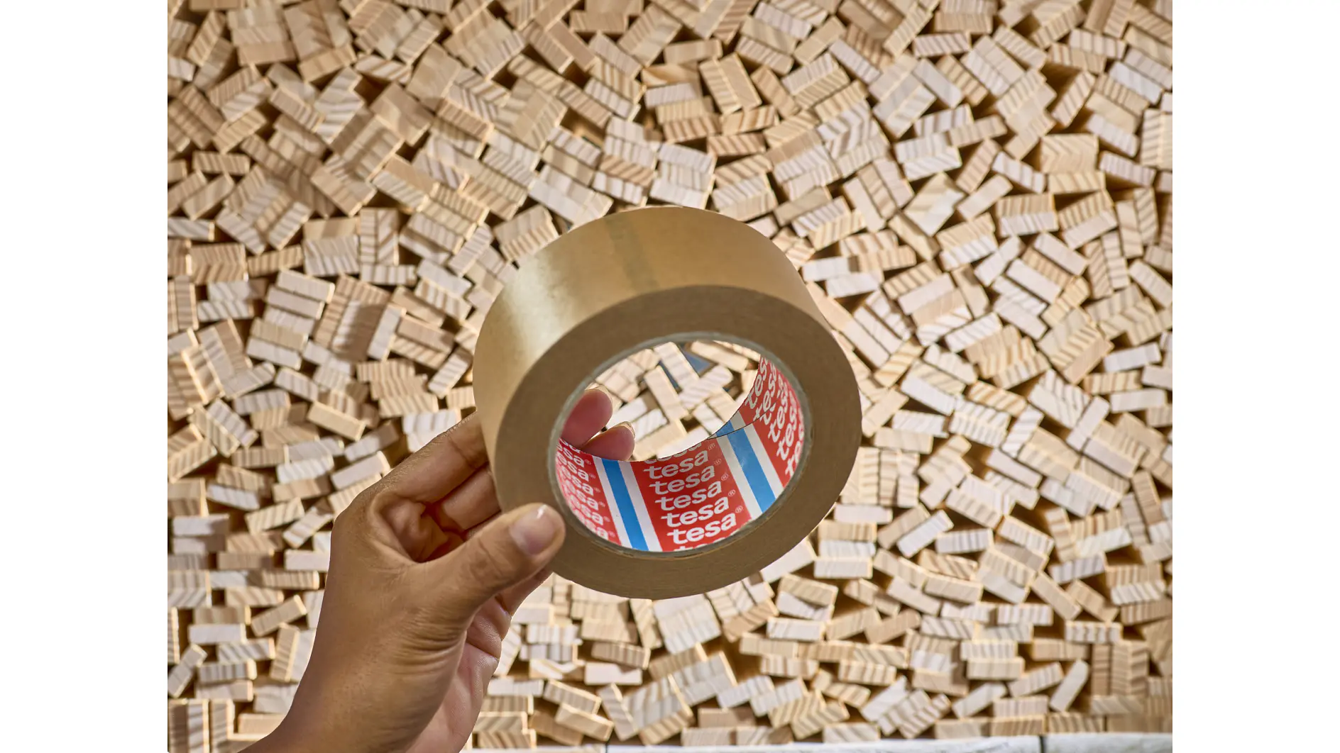 A hand holds a roll of brown tesa tape in front of a background filled with numerous small wooden blocks. (This text has been generated by AI)