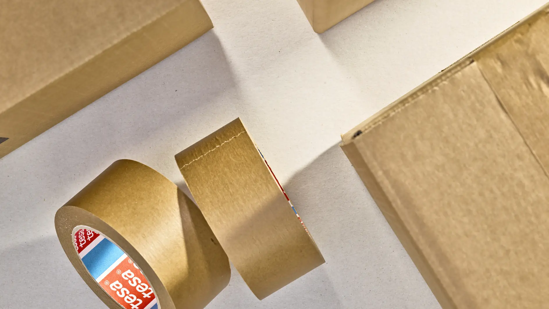 A roll of brown packing tesa tape is partially unrolled on a flat surface alongside closed cardboard boxes. The tesa tape label displays a red, white, and blue color scheme with text. The scene is lit from above, casting shadows.