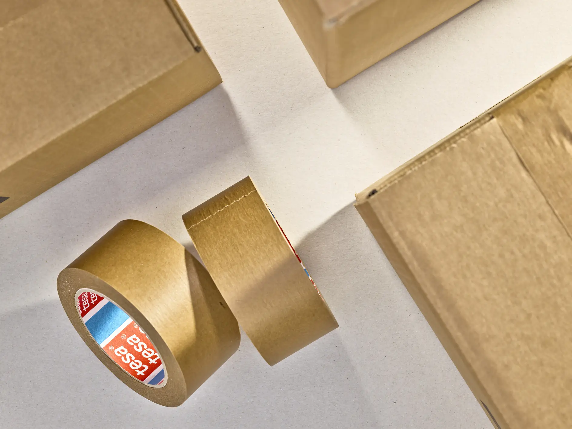 A roll of brown packing tesa tape is partially unrolled on a flat surface alongside closed cardboard boxes. The tesa tape label displays a red, white, and blue color scheme with text. The scene is lit from above, casting shadows.