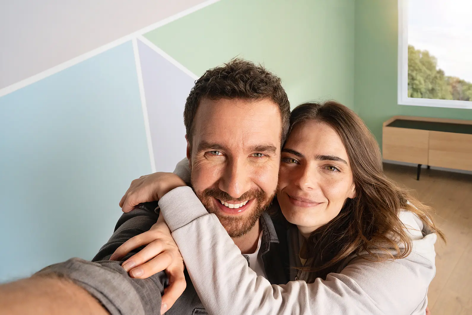 A man and a woman are smiling and taking a selfie in a room with light green walls and a geometric design. The woman has her arms around the mans shoulders. There is a window showing some greenery and a wooden cabinet in the background, adorned with tesa tape securing lightweight decorations to add flair to their space. (This text has been generated by AI)